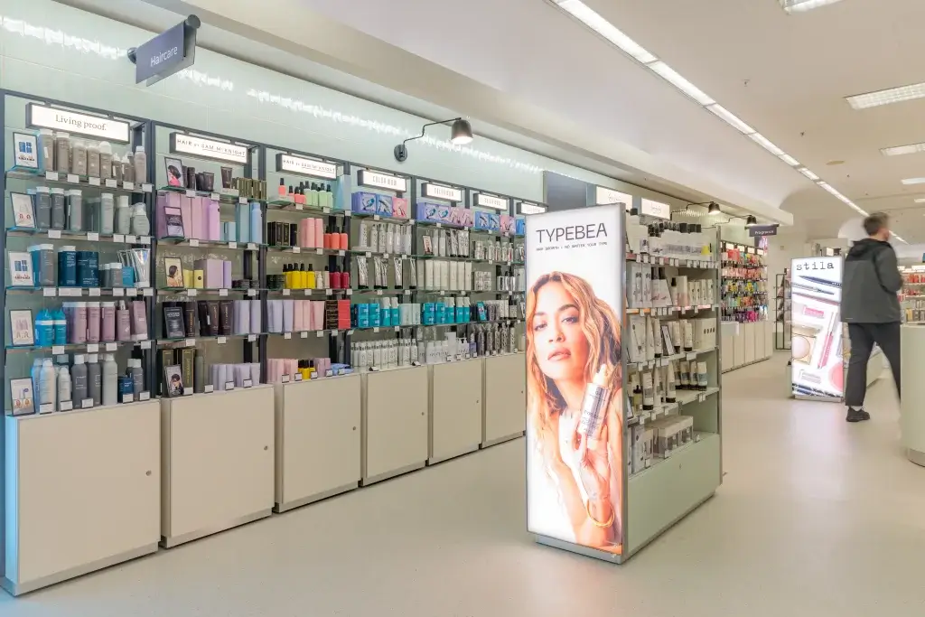 Cosmetics aisle with shelves of haircare products and an illuminated TYPEBEA display of a model holding a bottle.