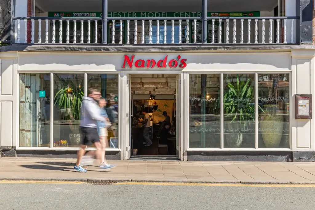 A bustling Nando's restaurant entrance with patrons inside, featuring a bright sign and green plants in the window.