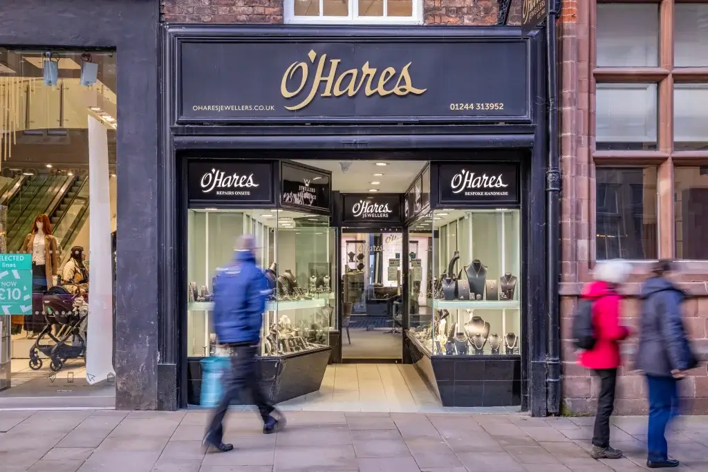 A blurred figure walks past the O'Hares Jewellers shop, showcasing elegant jewelry displays under a sophisticated black and gold sign.