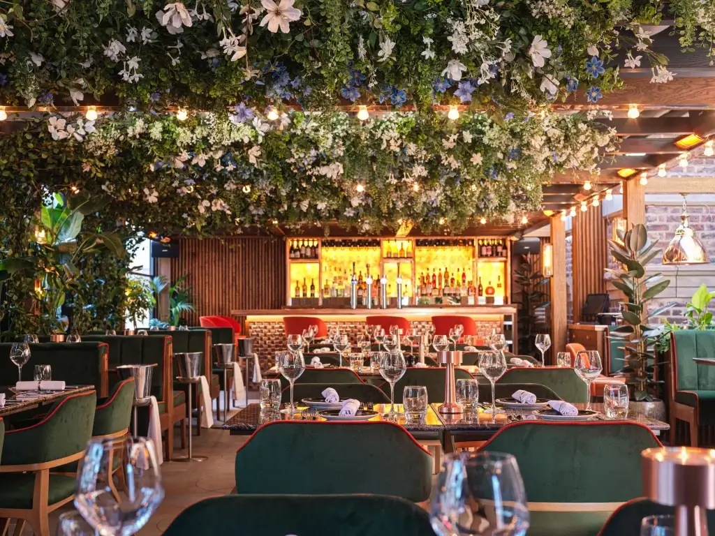 A vibrant restaurant interior adorned with lush greenery and flowers, featuring elegant seating and a stylish bar in the background.