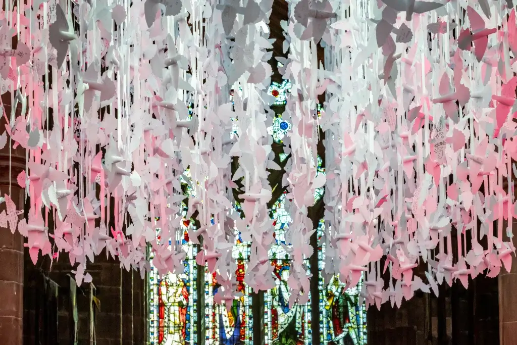 A cascade of paper dove shapes hung on ribbons in a grand cathedral with stained glass windows, adorned with handwritten messages, creating a poignant display of remembrance and hope