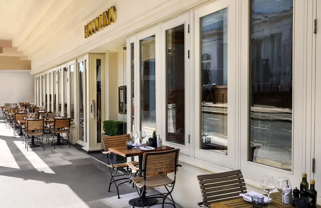 Outdoor dining area of Piccolino restaurant, featuring wooden tables and chairs, surrounded by large glass windows and a sunny ambiance.