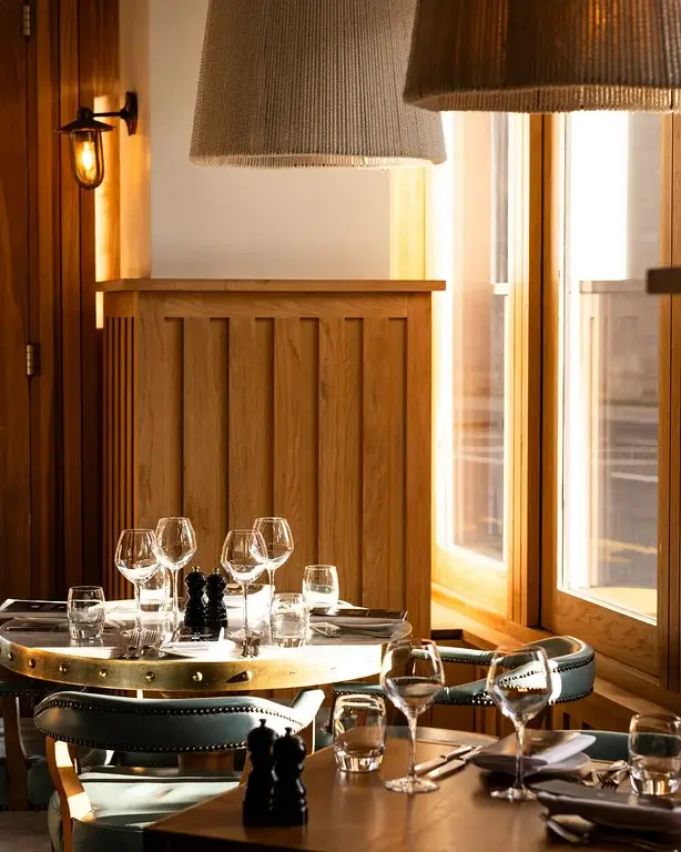 A cozy restaurant setting featuring a well-set table with glasses, cutlery, and soft lighting from large lamps and windows.
