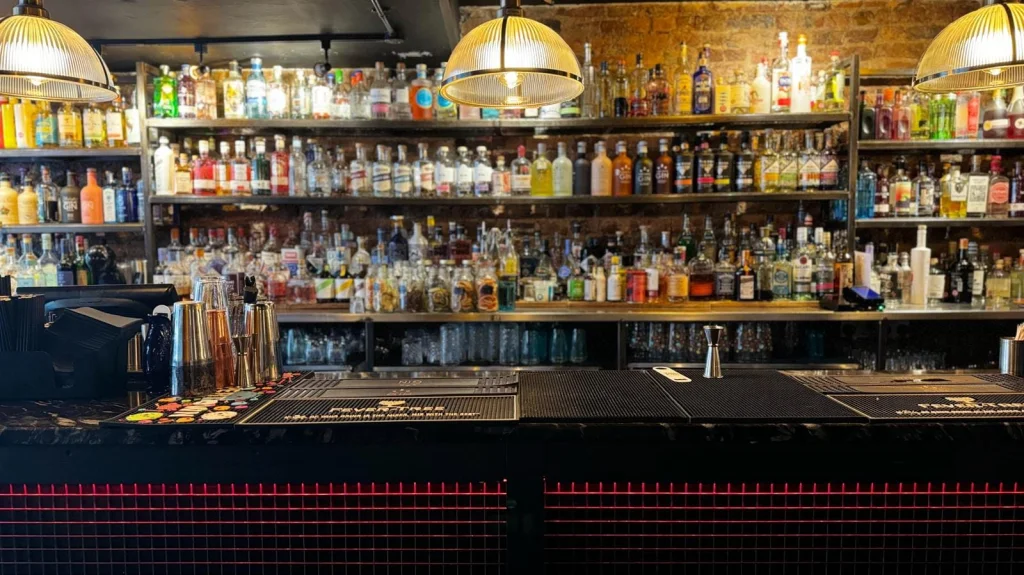 Long bar counter with metal shakers and jigger, shelves of assorted liquor bottles against exposed brick, warm pendant lights.