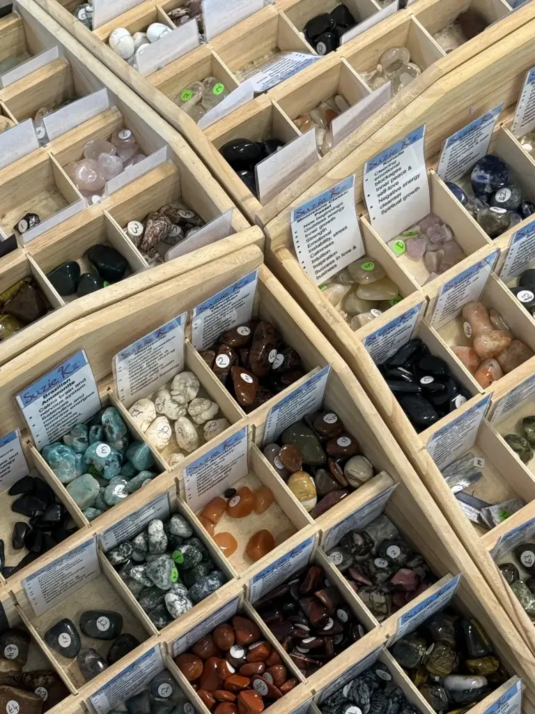 A top-down view of organized wooden boxes filled with various colorful gemstones, each labeled with names and prices.