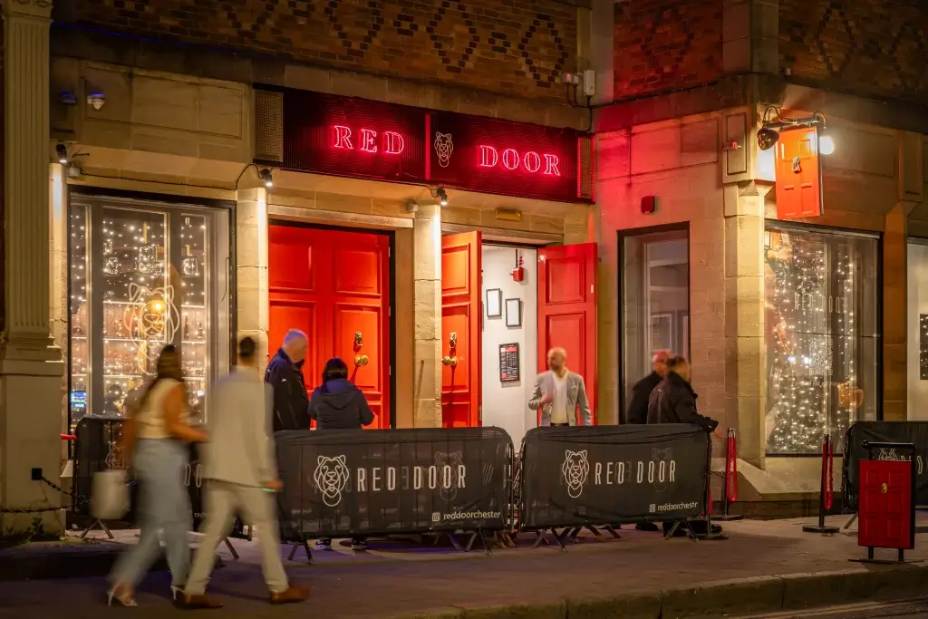 Exterior of Red Door bar with bright red doors and signage, illuminated by neon lights. People gather outside, adding a lively atmosphere.