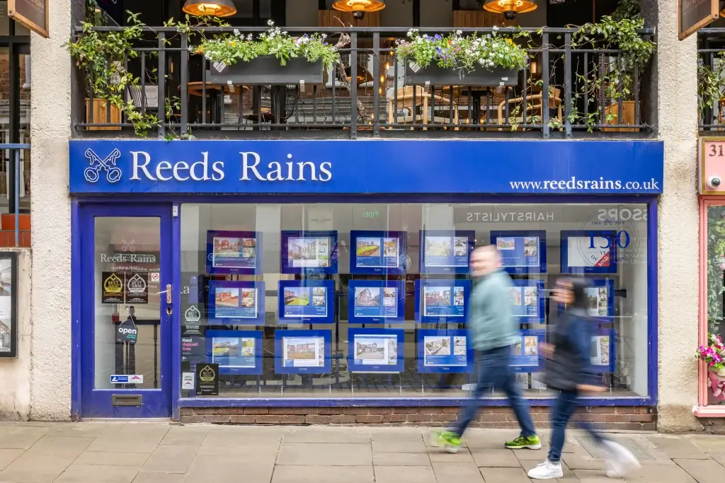 Storefront of Reeds Rains, featuring rental listings in framed displays and "open" signage, against a blue background.