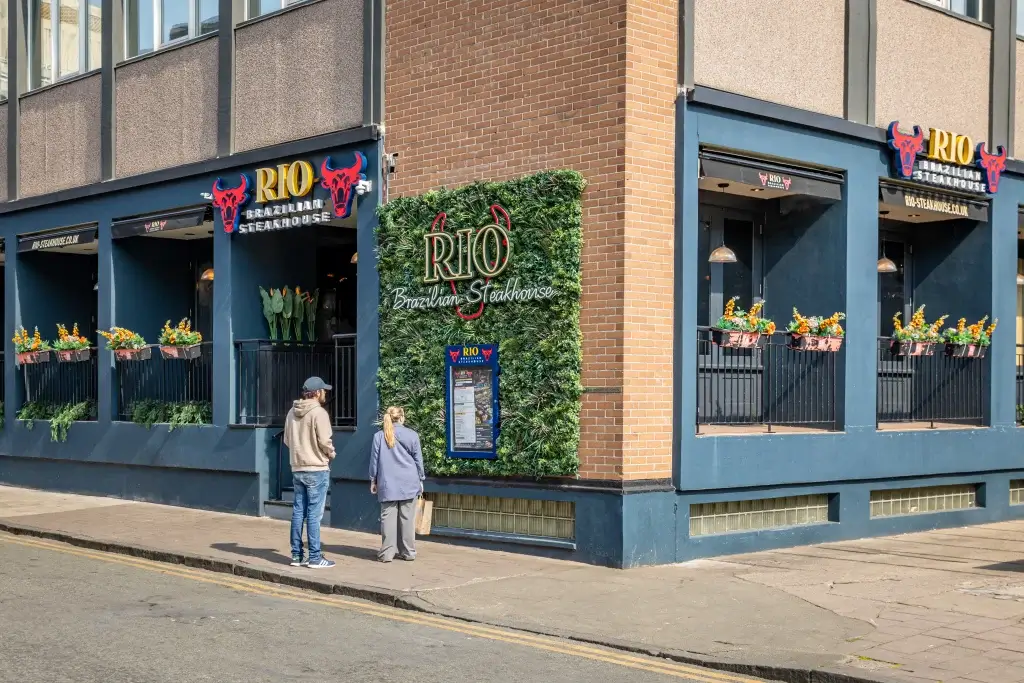 Two customers stand outside Rio Brazilian Steakhouse, admiring the vibrant exterior adorned with greenery and the restaurant's illuminated sign.