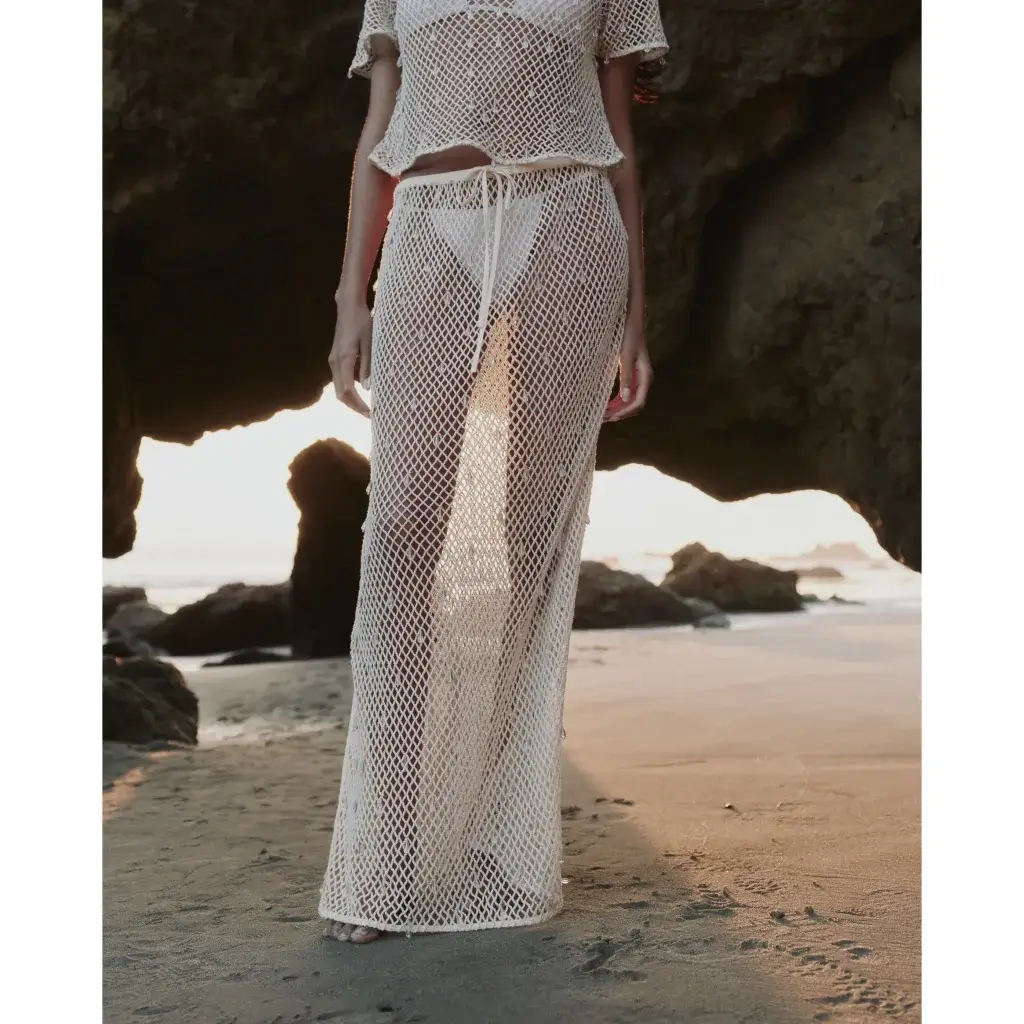 Person wearing a white crochet mesh skirt and bikini bottoms, standing barefoot on a sandy beach at sunset by a rock arch.