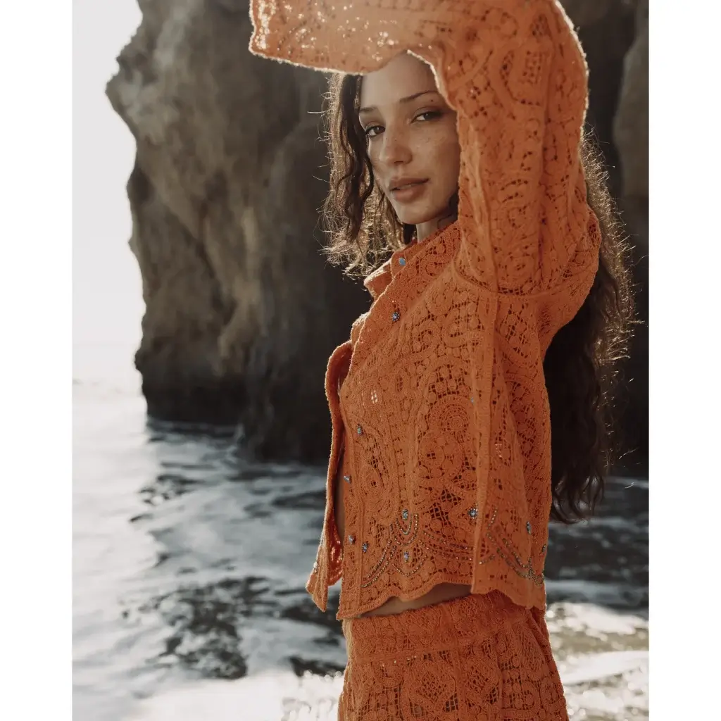 Woman at a rocky shore wearing an orange crocheted top and skirt, arm raised, sunlit hair and freckles.