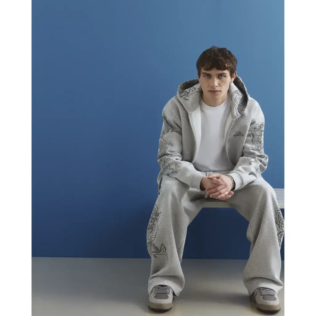 Young man sitting against a blue wall wearing a grey hoodie and matching sweatpants with black dragon prints, hands clasped.