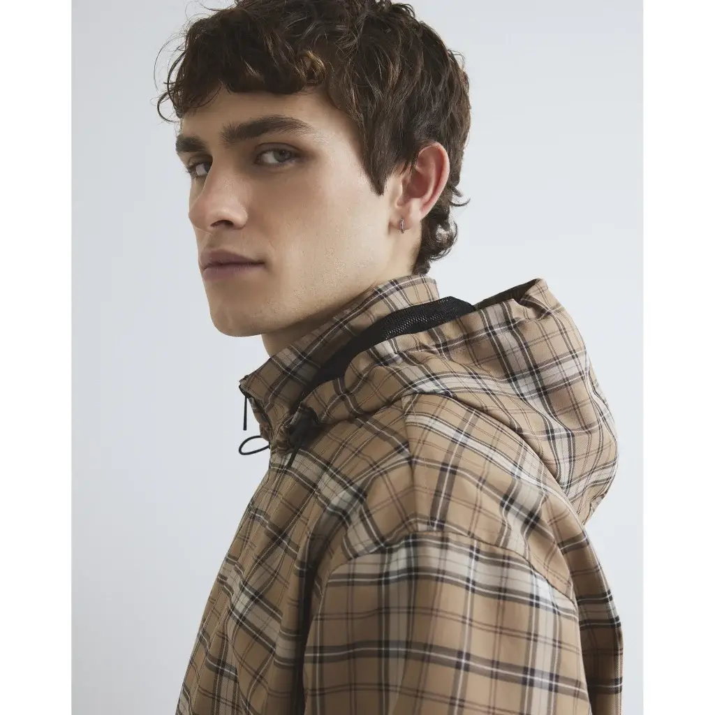 Young man in beige plaid hooded jacket seen in three-quarter profile with small hoop earring and neutral expression.