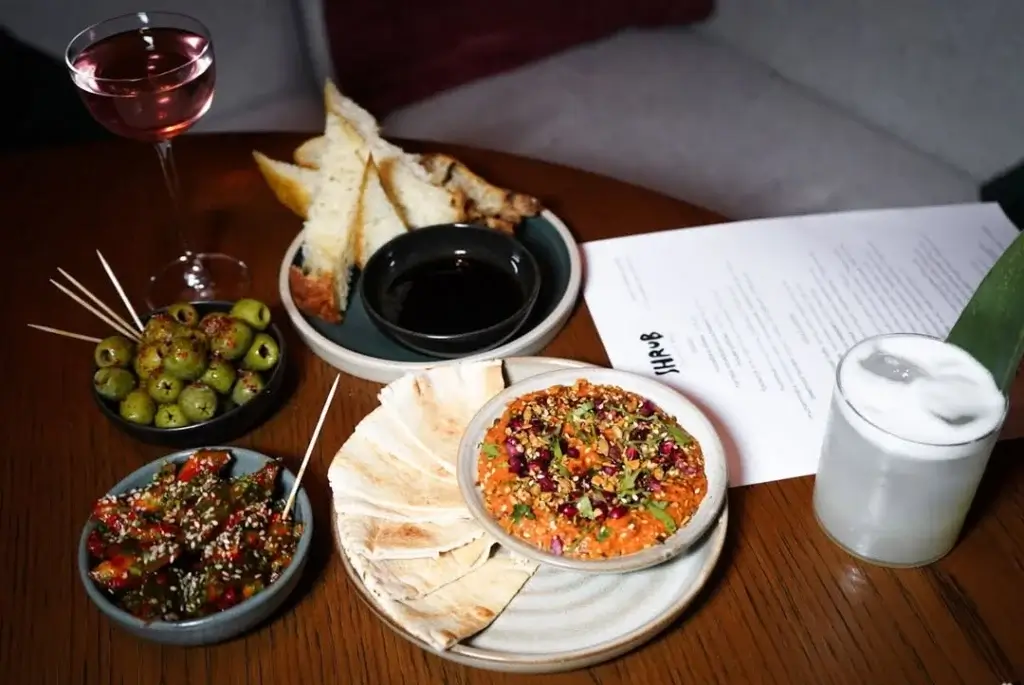 A stylish table setting featuring a cocktail, olives, dips, bread, and pita alongside a menu, all invitingly presented.