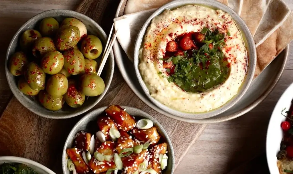 A spread of green olives with spices, creamy hummus topped with herbs, and a sesame-topped dish, accompanied by pita bread.