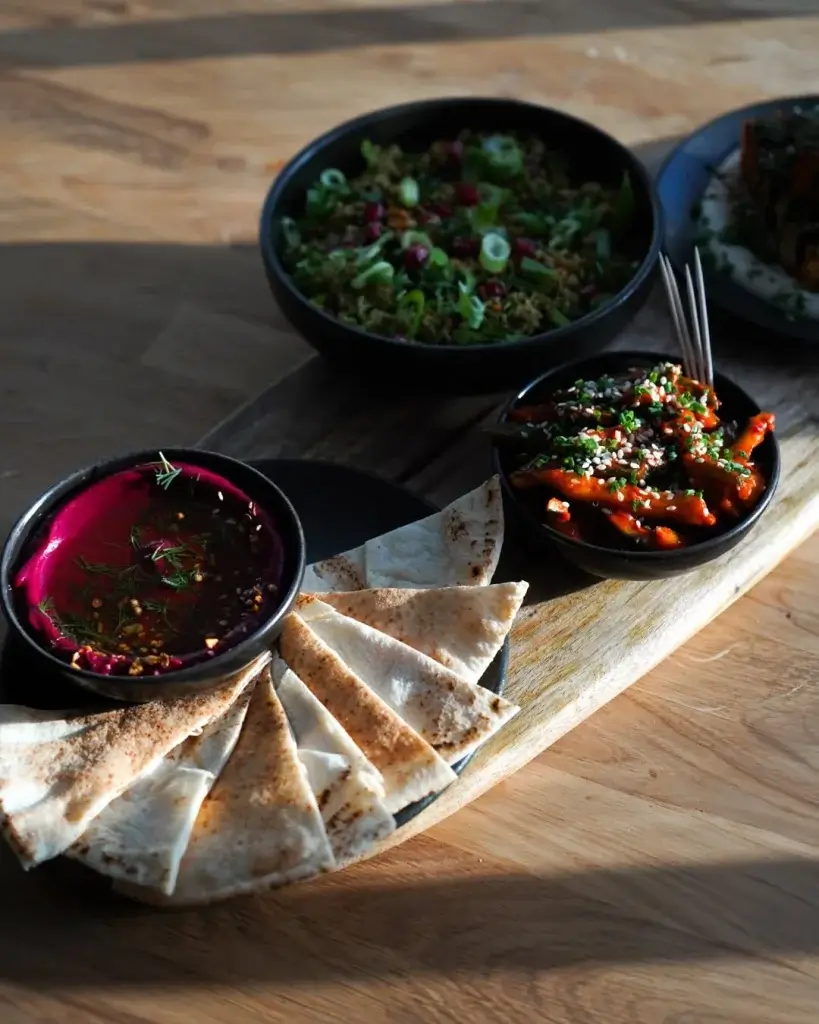 A wooden platter holds colorful bowls of vibrant dip, salad, and saucy dish, accompanied by freshly baked flatbreads.