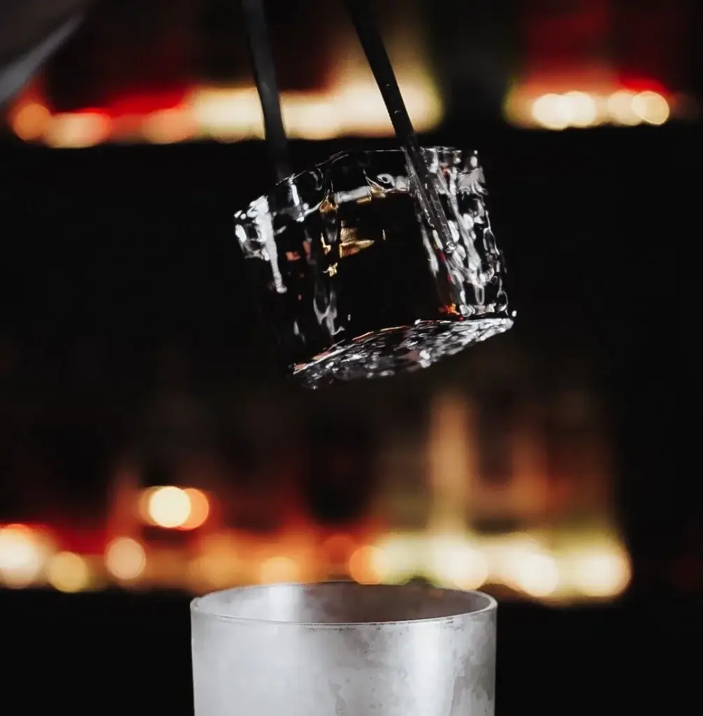 A clear ice cube is suspended above a glass, ready to be dropped, with a blurred bar background illuminated by soft lights.