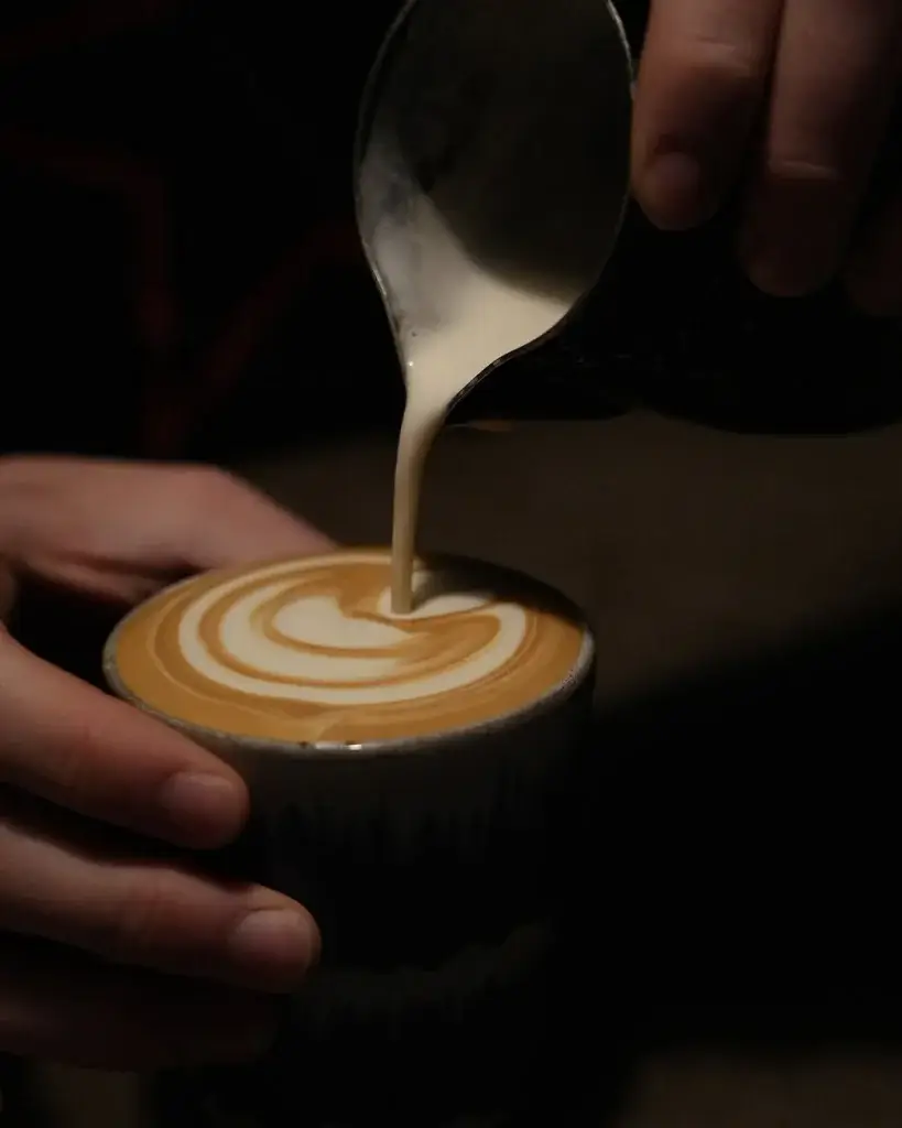 A hand pours steamed oat milk into a cup of coffee, creating a beautiful swirl pattern on the frothy surface.
