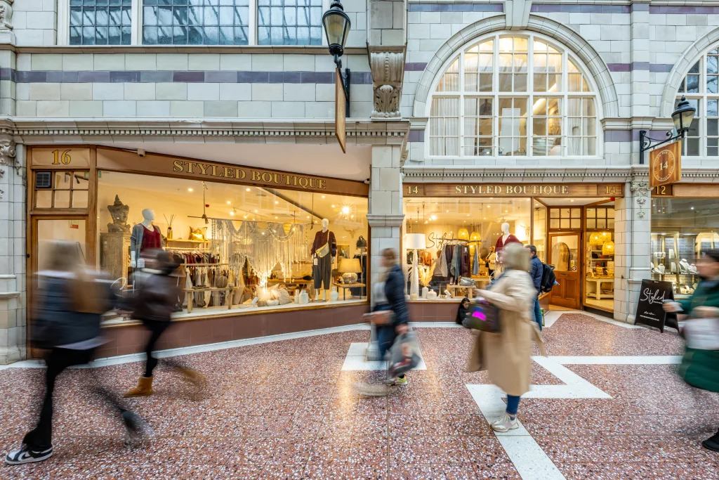 Two stylish boutique storefronts with mannequins displaying clothing, set in a charming indoor shopping arcade. Shoppers walk by.