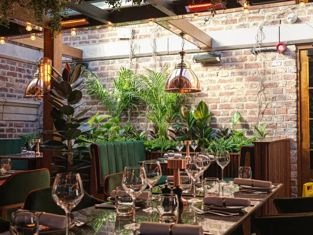 A stylish restaurant interior with green upholstery, lush plants, and warm lighting, featuring a glass-topped dining table set for guests.