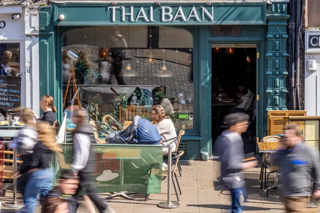 A vibrant Thai restaurant named "Thai Baan," featuring outdoor seating, plants, and busy pedestrians in a sunny urban setting.