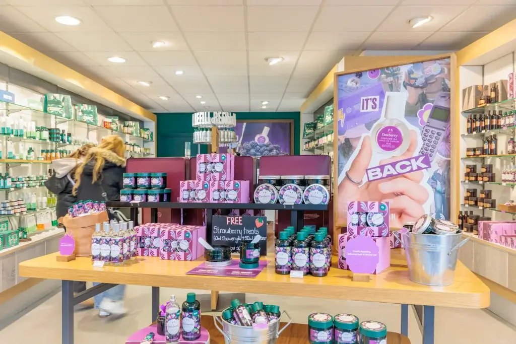 Table display of purple-packaged shower gels, creams and tubs with a large "It's Back" poster in a bright cosmetics shop