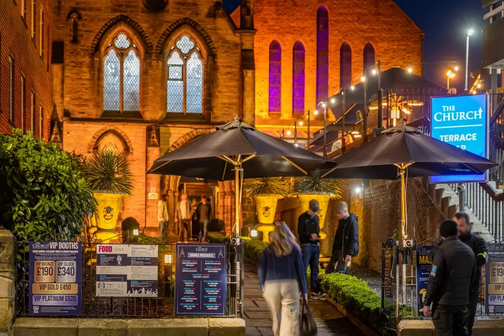 A lively terrace bar at night, featuring umbrellas, a bright sign, and guests enjoying the ambiance amid warm lighting and greenery.