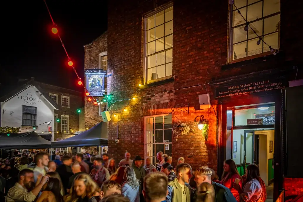 A lively outdoor gathering at night with warm lights and a historic building, filled with people enjoying the atmosphere.