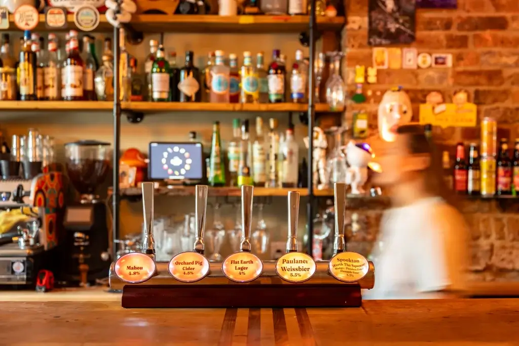 A wooden beer tap display with five labels in front of a lively bar adorned with various spirits and a blurred bartender.