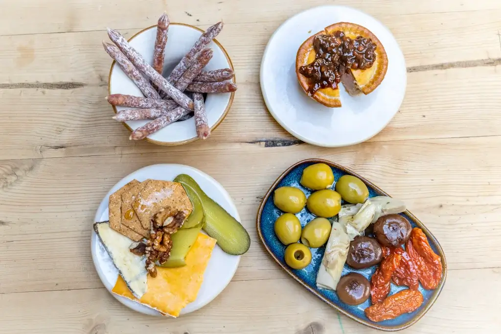 A rustic spread featuring plates of cured meats, cheese, olives, pickles, and a sweet pastry on a wooden table.