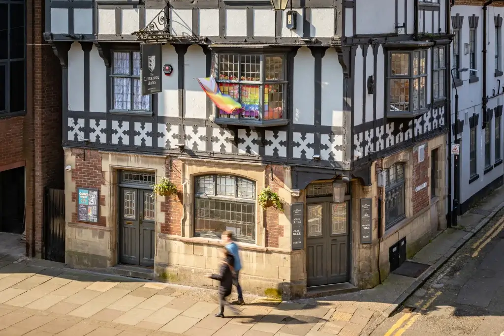 The Liverpool Arms pub features Tudor-style architecture with a rainbow flag displayed above the entrance as two people walk by.