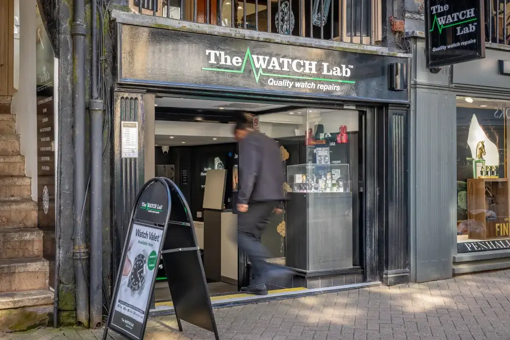 A blurred figure exits The Watch Lab, a watch repair shop showcasing various timepieces in the window display.