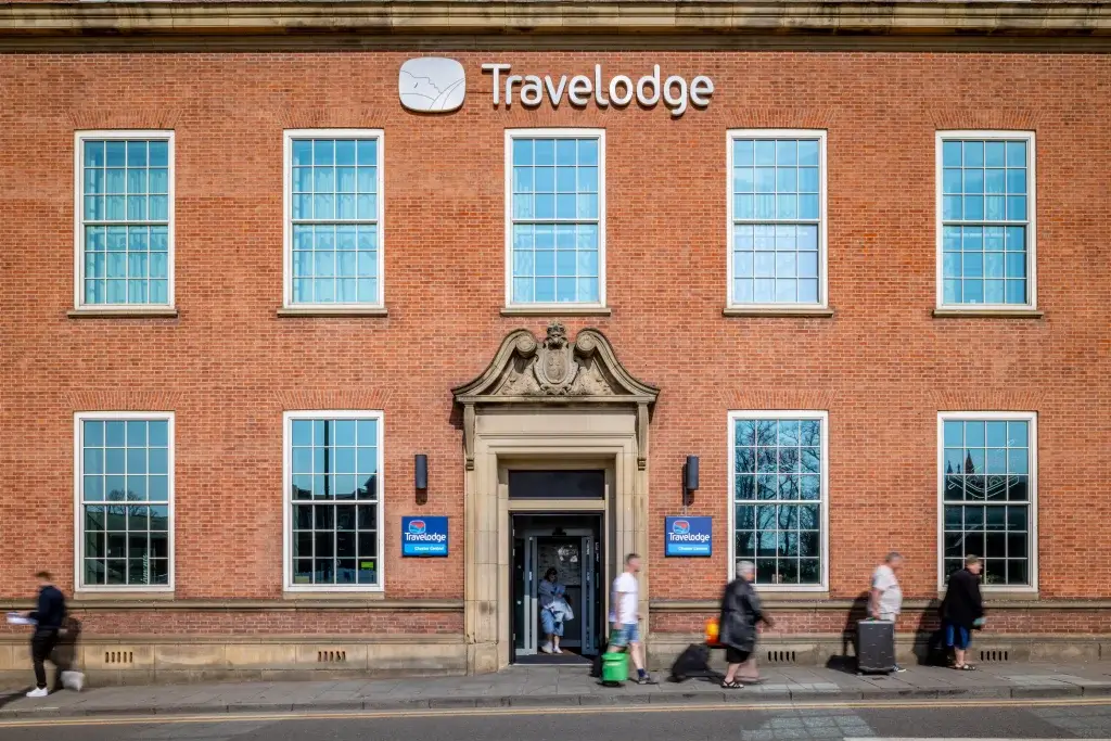 Exterior view of a Travelodge hotel, featuring a brick facade and large windows, with guests entering and exiting.