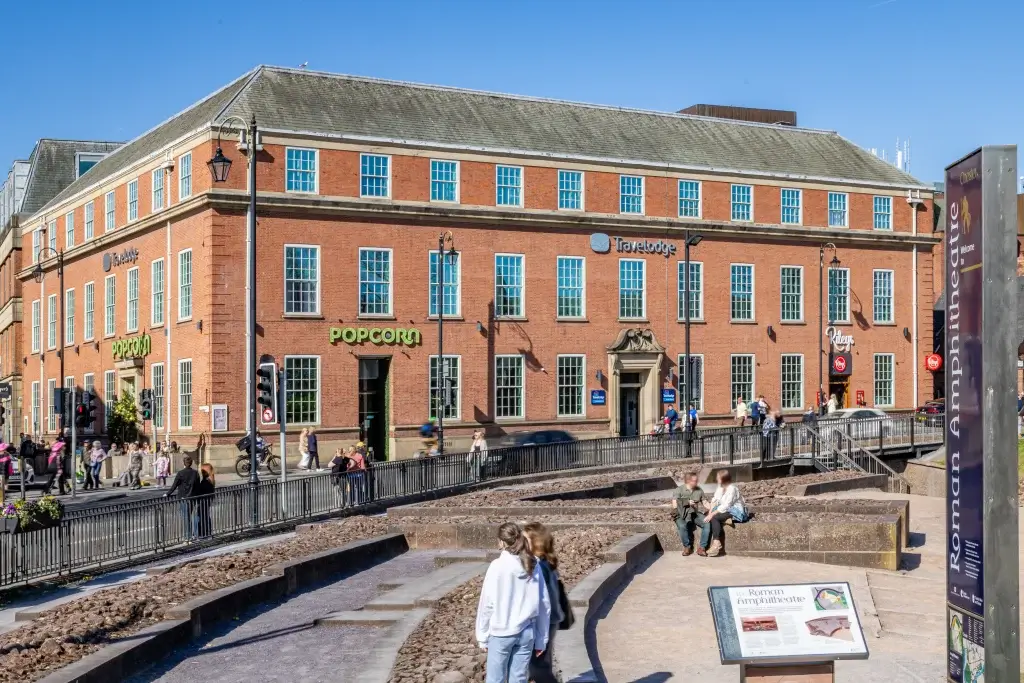 A busy city street features a red brick building with 'Travelodge' and 'Popcorn' signage, surrounded by pedestrians and traffic lights.