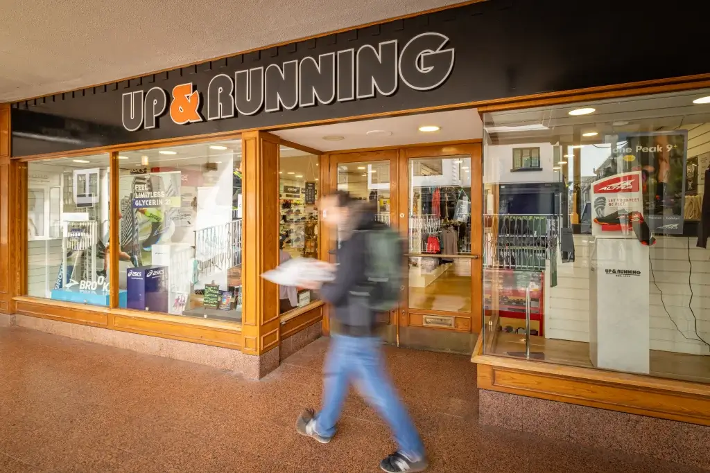 A blurred figure walks by the entrance of the "UP & RUNNING" store, showcasing athletic gear and footwear through the window.