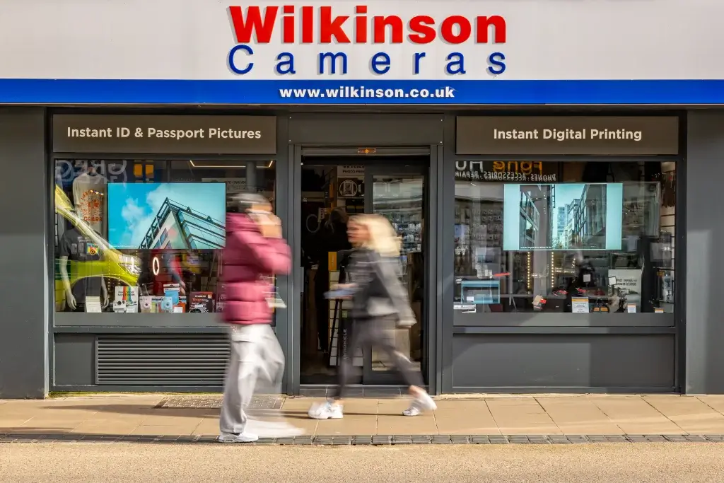 A storefront labeled "Wilkinson Cameras" featuring two screens displaying photos, with people walking by and a digital printing sign above.