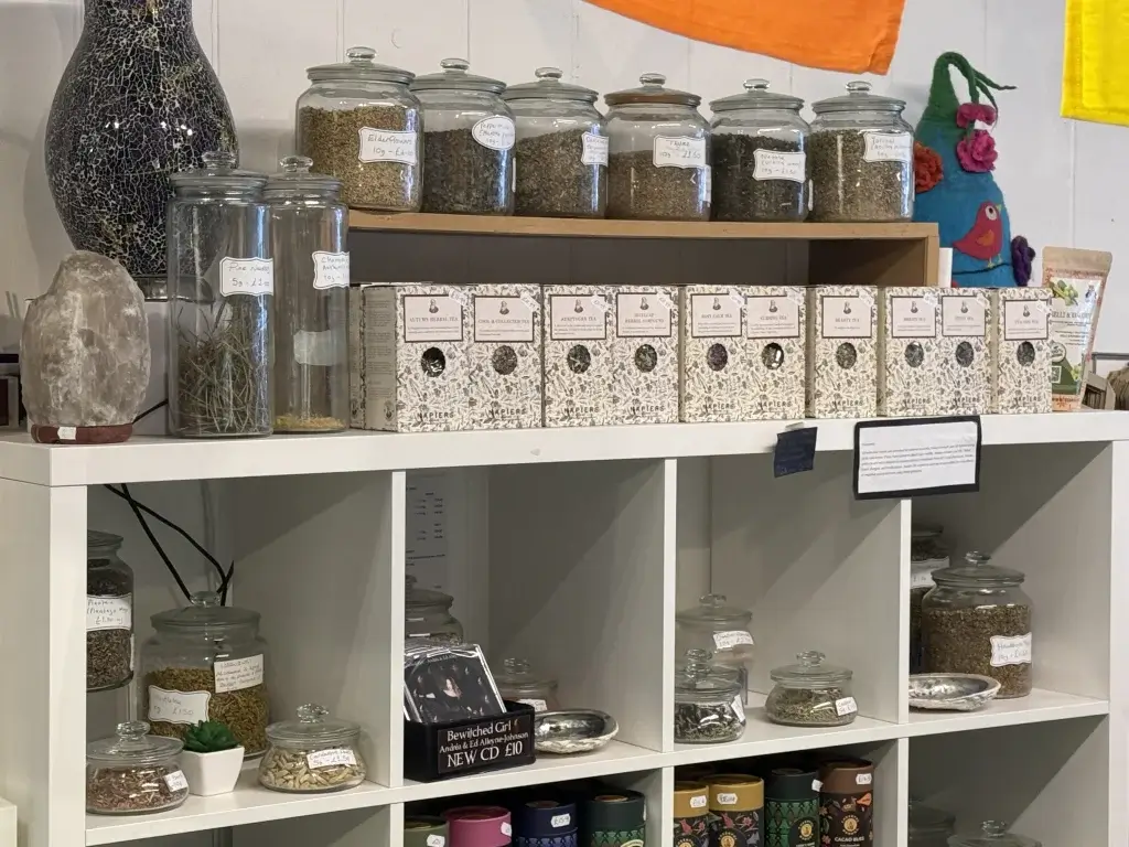 A vibrant herbal shop display featuring jars of herbs, assorted tea boxes, and a decorative stone on a white shelf, with colorful fabrics above.