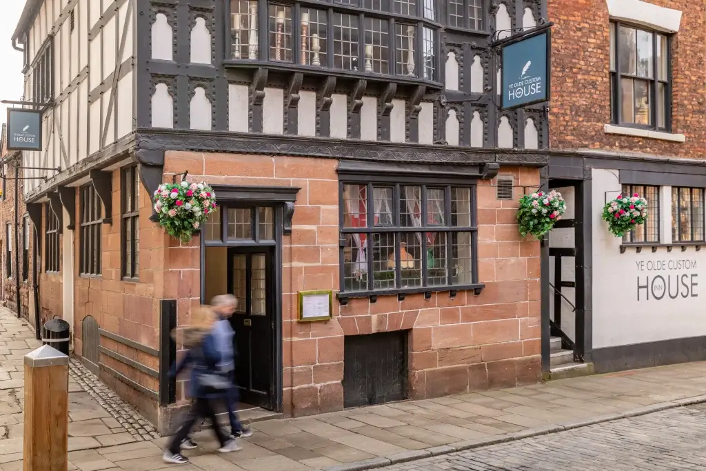 Historic building with black timber framing and large windows, featuring hanging flower baskets and a sign for "Ye Olde Custom House."