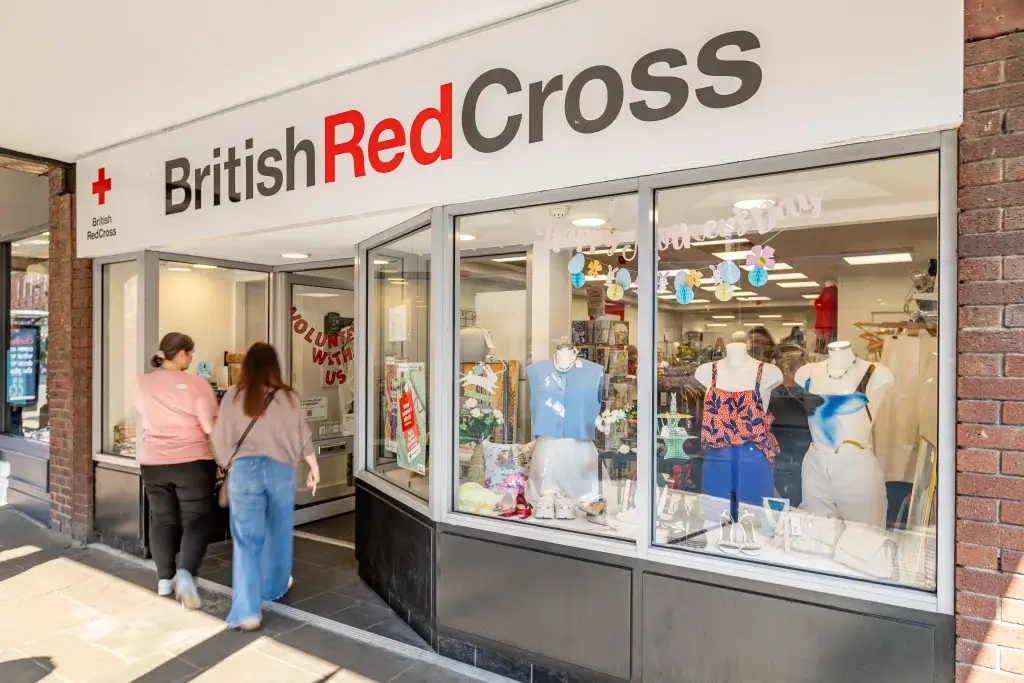 Two women enter the British Red Cross shop, showcasing colorful clothing and decor in the window display celebrating Mother’s Day.