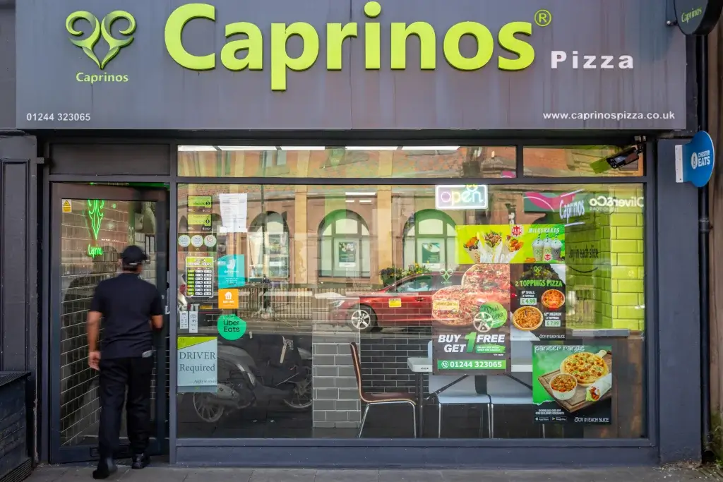 Exterior view of Caprinos Pizza restaurant, featuring a vibrant sign, 'Open' neon light, and promotional offers displayed in the window.