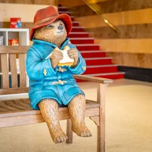 A statue of Paddington Bear sitting on a bench infront of bookcase on and stairwells either side