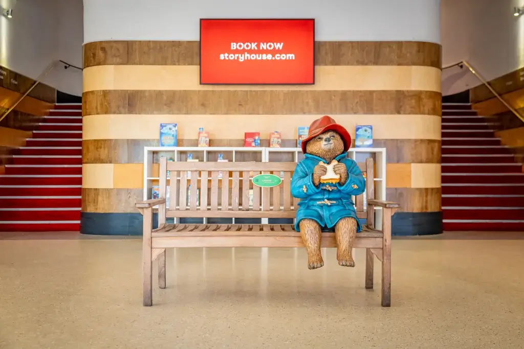 A statue of Paddington Bear sitting on a bench infront of a tv with Storyhouse on and stairwells either side