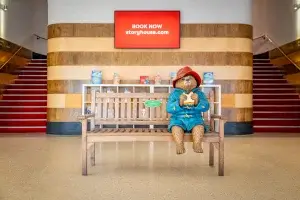 A statue of Paddington Bear sitting on a bench infront of a tv with Storyhouse on and stairwells either side