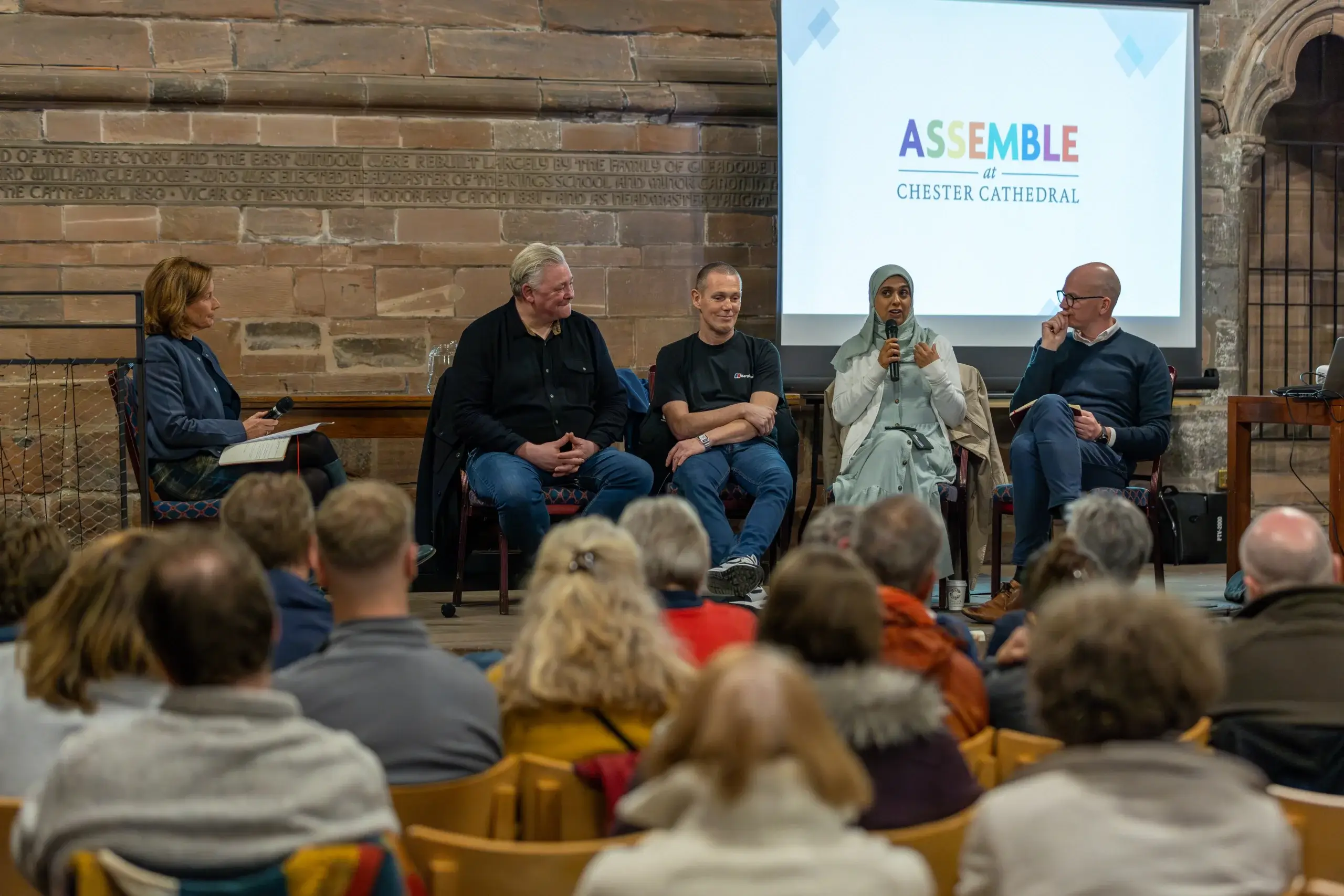A panel discussion at Chester Cathedral, featuring speakers on stage, with an audience engaged in the event. A colorful banner reads "ASSEMBLE."