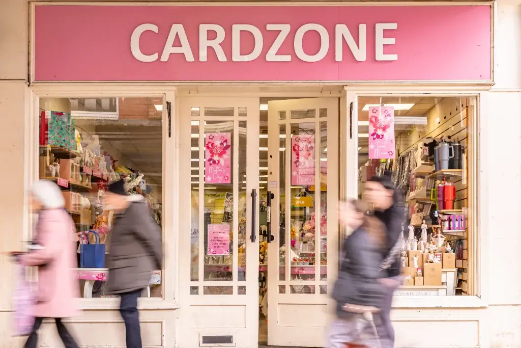 Entrance of CARDZONE store with pink signage and blurred pedestrians walking by, showcasing a colorful display of gifts inside.