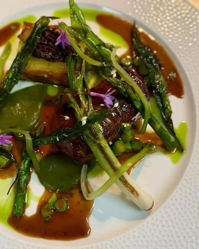 A gourmet dish featuring sautéed greens, grilled meat, vibrant sauces, and edible flowers, artistically arranged on a white plate.