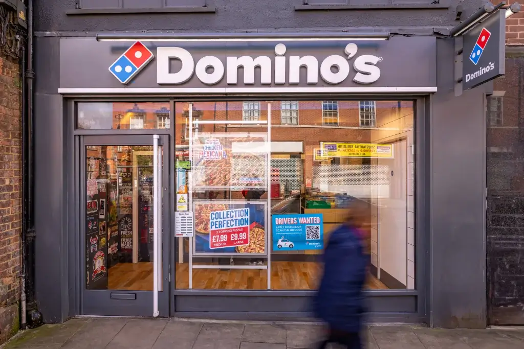 Domino’s storefront with promotions displayed, featuring a blurred pedestrian passing by, creating a lively urban scene.