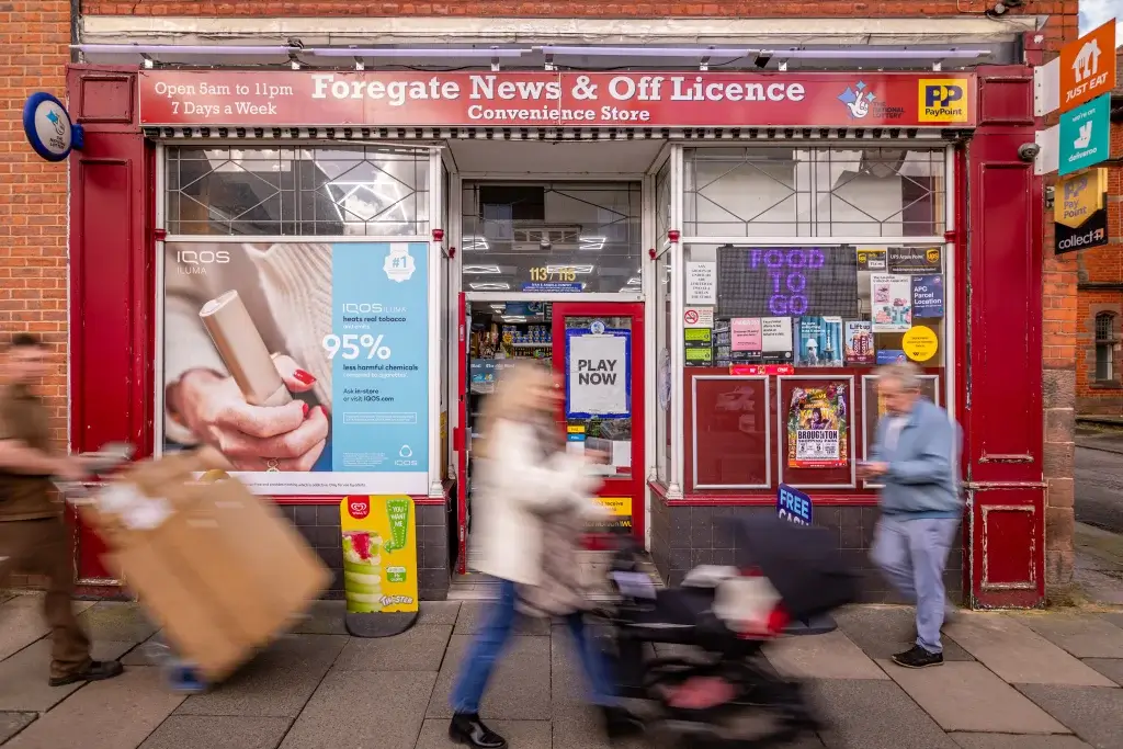 Facade of Foregate News & Off Licence convenience store with a colorful sign, advertisements, and pedestrians passing by.