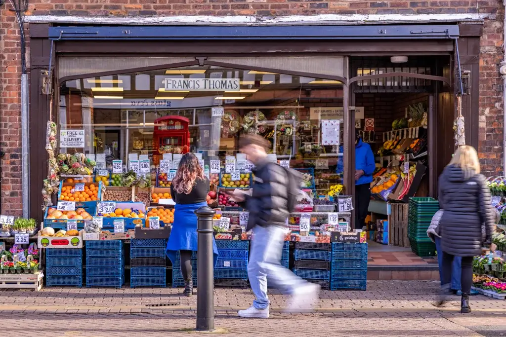 A colorful fruit and vegetable shop with customers browsing outside, displaying fresh produce and seasonal flowers.