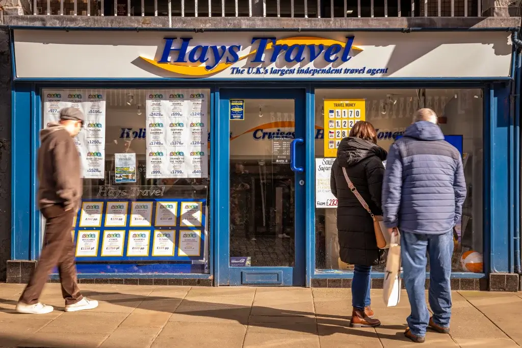 Hays Travel storefront with travel offers displayed in the window, while two individuals examine the advertisements.