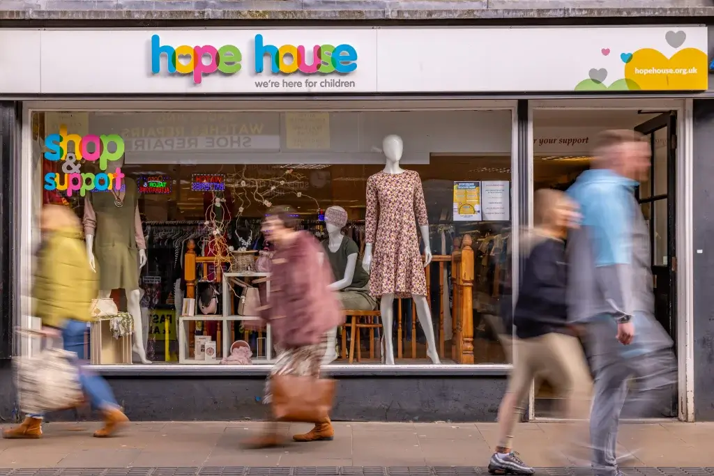 A busy storefront of Hope House charity shop featuring colorful signage, a clothing display, and shoppers passing by on the street.
